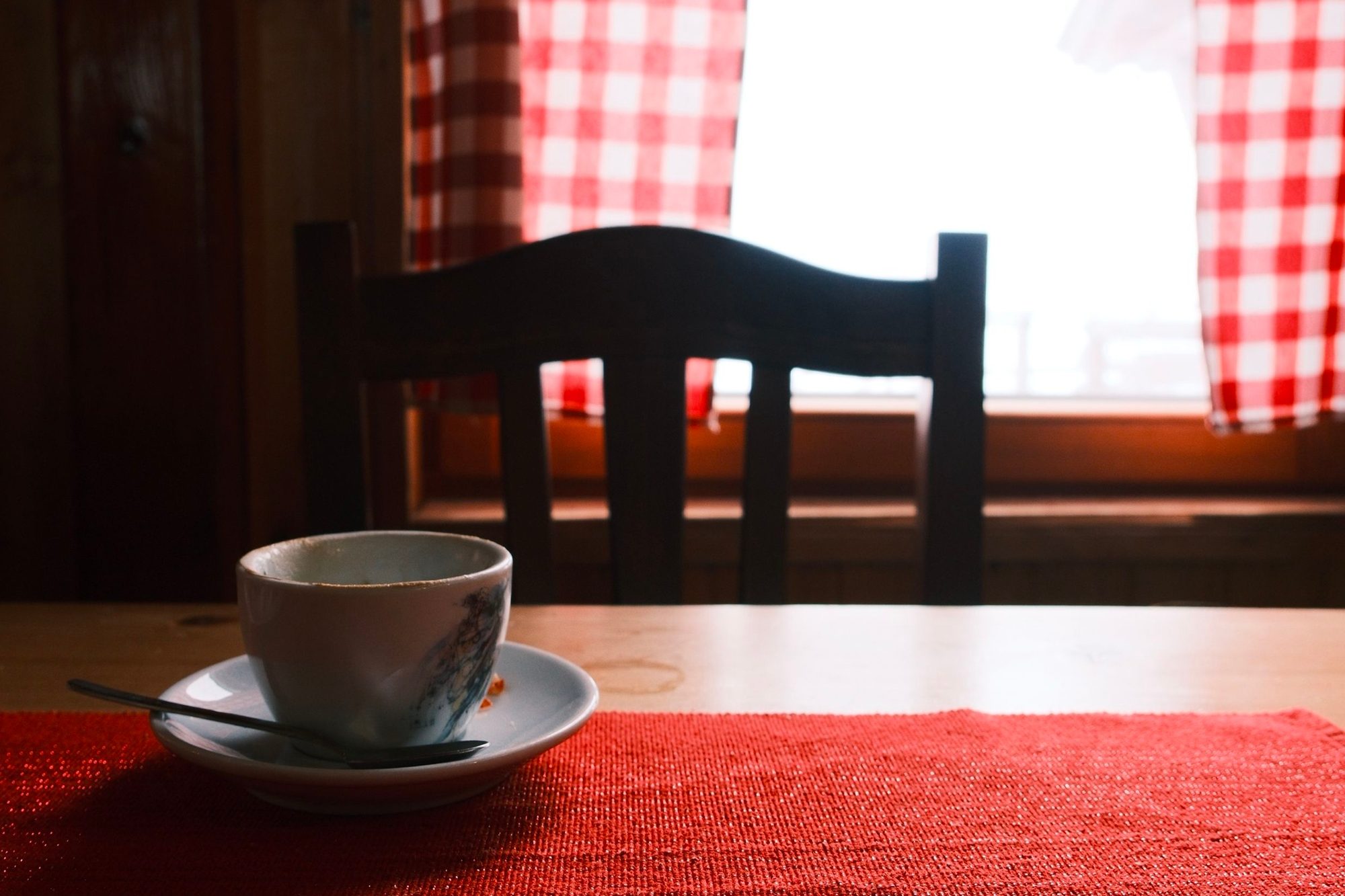An empty wooden chair at a table by a curtained window, with a used cup and saucer left on the table, as though someone has just got up and stepped out of the room.