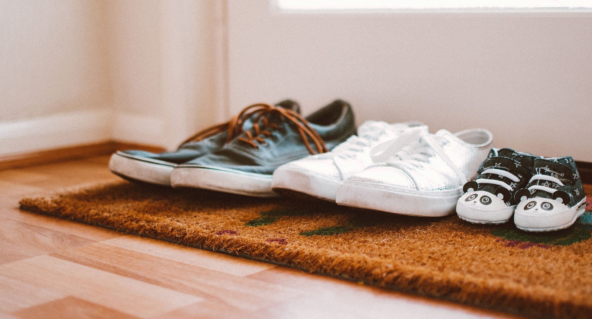 Three pairs of shoes lined up on a coir mat by a front door — two adult pairs and, at the end, a pair of baby shoes.